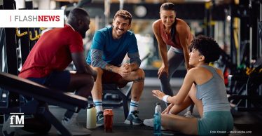 Vier junge Erwachsene machen im Fitnessstudio eine Trainingspause und sitzen im Kreis auf dem Boden; sie lachen und reden miteinander.