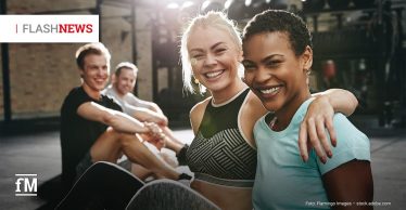 Zwei Frauen sitzen lächelnd im Fitnessstudio, eine legt den Arm um die andere, im Hintergrund zwei weitere Personen auf Trainingsmatten, Schriftzug „FLASH NEWS“ oben links.