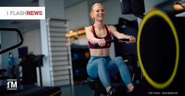 Eine sportlich gekleidete junge Frau trainiert in einem modernen Fitnessstudio an einem Rudergerät und lächelt dabei in Richtung Monitor.