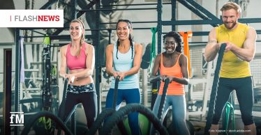 Vier Personen, zwei Frauen links, eine Frau und ein Mann rechts, trainieren lachend gemeinsam mit schweren Battle Ropes in einem Fitnessstudio.