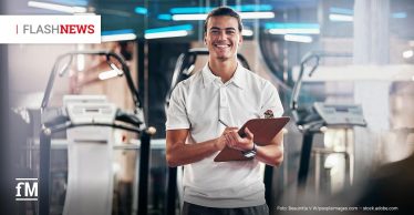 Ein junger Mann in weißem Polohemd in einem Fitnessstudio mit Klemmbrett und Stift; im Hintergrund Laufbänder und Fitnessgeräte.