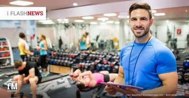 Mann im blauen Shirt mit Klemmbrett steht in Fitnessstudio, im Hintergrund trainieren mehrere Personen mit Hanteln.