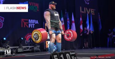 Ein muskulöser Strongman-Athlet (Hafthor Björnsson) hebt auf einer Bühne eine Langhantel mit 510 kg Gewicht; er trägt Sportkleidung, Gewichthebergürtel und steht bei seinem Weltrekord vor einer Reihe internationaler Flaggen.
