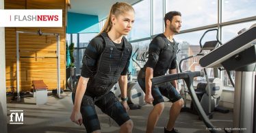 Zwei Personen, eine Frau und ein Mann, trainieren in einem Fitnessstudio mit Elektro-Muskelstimulationsanzügen; sie stehen in leichter Kniebeuge neben einem Laufband