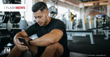 Ein Mann in Sportkleidung sitzt nach dem Training in einem Fitnessstudio auf dem Boden und schaut konzentriert auf sein Smartphone. Im Hintergrund sind Hanteln und Trainingsgeräte zu sehen.