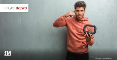 Ein junger Mann in Sportkleidung steht vor einer grauen Wand, trägt einen rostroten Hoodie und schwarze Shorts. In der rechten Hand hält er eine kleine Kettlebell, mit der linken Hand zeigt er nachdenklich auf seinen Kopf.