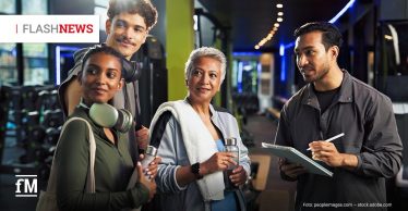 Vier Personen in Sportkleidung stehen mit fragenden Gesichtern in einem modernen Fitnessstudio und hören einem Trainer mit Klemmbrett aufmerksam zu.