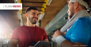 Ein junger Personal Trainer im roten Shirt hält ein Tablet und lacht, während er mit einem älteren Mann im blauen Shirt und Stirnband spricht; sie stehen in einem Fitnessstudio.