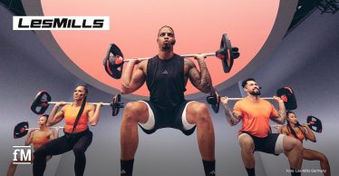 Mehrere Trainierende führen in einem Studio gemeinsam Kniebeugen mit Langhanteln vor einer beleuchteten Trainingsfläche aus.