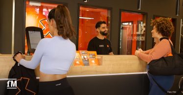 Zwei Frauen stehen an der Rezeption des Fitnessstudios FitX, eine checkt sich an einem Tablet ein, die andere spricht lächelnd mit einem Mitarbeiter hinter dem Tresen. Im Hintergrund steht in großen weißen Buchstaben „FOR ALL OF US“.