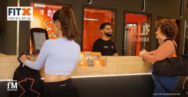Zwei Frauen und ein Mitarbeiter stehen am Empfang eines FitX-Studios.