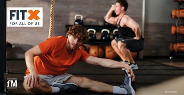 Zwei Trainierende in einem FitX Studio: vorne ein Mann im orangefarbenen Shirt beim Dehnen am Boden, hinten ein Mann bei einer Kniebeuge mit Gewichten.