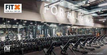 Moderner Freihantelbereich in einem FitX-Studio mit zahlreichen Hanteln, Trainingsbänken und Spiegelwänden. An der Wand der Slogan 'FOR ALL OF US'.