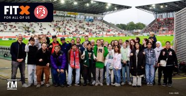 Eine große, bunt gemischte Gruppe junger Menschen posiert lächelnd am Spielfeldrand eines Stadions. Im Hintergrund sind die Tribünen mit einigen Zuschauern zu sehen. Oben links im Bild ist das Logo von FitX und Rot-Weiss Essen zu sehen.