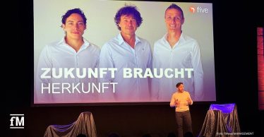 Bühne mit Vortrag, vorn ein Sprecher, dahinter große Leinwand mit drei Personen, five Logo und Schriftzug 'Zukunft braucht Herkunft', Publikum im Vordergrund.