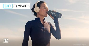 Eine sportliche Frau trägt Kopfhörer und trinkt aus einer schwarzen Flasche mit der Aufschrift 'Fitness Studio'. Sie steht draußen bei Tageslicht. Oben links steht das Logo 'GIFT CAMPAIGN'.