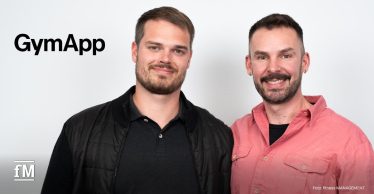 Zwei Männer stehen nebeneinander vor einer weißen Wand. Links trägt der Mann (Robin Lausch) eine schwarze Jacke und ein dunkles Poloshirt, rechts trägt der Mann (Janosch Marx) ein rosafarbenes Hemd. Links oben ist das schwarze GymApp-Logo zu sehen.