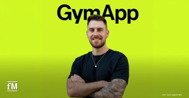 Mann mit tätowiertem rechten Arm steht mit verschränkten Armen vor neongelbem Hintergrund; über ihm steht groß der Schriftzug „GymApp“