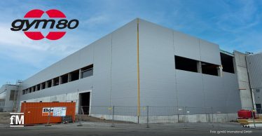 Auf dem Bild ist eine große, moderne Industriehalle im Bau zu sehen. Die Fassade ist hellgrau und besteht aus glatten, rechteckigen Paneelen. Links im Bild steht ein orangefarbener Baucontainer davor, umgeben von einem Bauzaun. Über dem Gebäude links oben ist das rote und schwarze gym80-Logo eingeblendet.