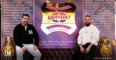 Zwei Männer sitzen auf einem braunen Ledersofa vor einem gym80-Logo mit goldenen Hantelscheiben links und rechts.