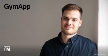 Porträt eines Mannes im dunklen Hemd (Robin Lausch), der an einer weißen Wand steht; links oben steht das Logo 'GymApp'.