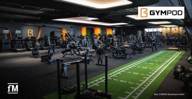 Innenraum eines Fitnessstudios mit schwarzem Boden und schwarzer Decke, orange beleuchteten Wandsegmenten im Hintergrund, links mehrere Laufbänder und Kraftgeräte in Reihen angeordnet, mittig Rudergeräte und Trainingsmaschinen, rechts eine grüne Kunstrasenfläche mit weißen Markierungen, im Vordergrund ein Schlitten mit Gewichtsscheiben, Blick aus leicht erhöhter Perspektive in den Raum