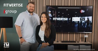 Zwei Personen stehen vor einem Bildschirm mit den Logos „FITVERTISE“ und „igroupTV“. Im Vordergrund befindet sich ein Holztisch. Hinter den Personen verläuft eine Wand mit vertikalen Holzleisten und einem großen Monitor.