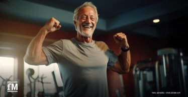 Älterer Mann mit grauem Haar, Bart und T-Shirt steht lachend im Fitnessstudio und zeigt seine Muskeln. Im Hintergrund stehen Fitnessgeräte.