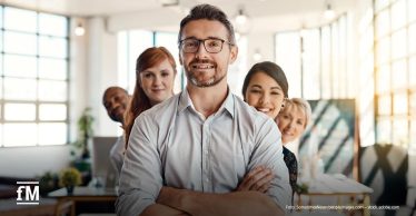 Fünf lächelnde Personen unterschiedlicher Herkunft stehen eng beieinander in einem hellen Büro, vorne ein Mann mit Brille und verschränkten Armen.