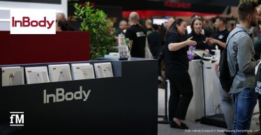 Im Vordergrund ein schwarzer Messestand mit weißem Schriftzug „InBody“ und Prospektfächern; dahinter mehrere Personen im Gespräch; oben links rotes InBody-Logo; Messehalle mit weiteren Ständen im Hintergrund.