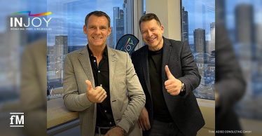 Zwei Männer in Business-Outfits stehen lächelnd vor einem Fenster mit Blick auf die Skyline, beide zeigen den Daumen hoch. Links oben das INJOY-Logo.