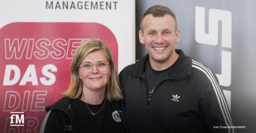 Zwei Personen stehen nebeneinander und lächeln in die Kamera. Links eine Frau mit schulterlangen blonden Haaren, Brille und schwarzer Jacke mit dem Logo „fM – fitness MANAGEMENT“. Rechts ein Mann in schwarzer Adidas-Sportjacke mit weißen Streifen. Im Hintergrund sind ein rotes Schild mit weißer Schrift „WISSEN DAS DIE“ sowie ein graues Banner mit weißen Buchstaben zu sehen.