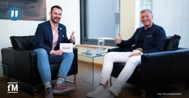 Zwei Männer sitzen sich in Sesseln gegenüber, beide zeigen mit einem Lächeln den Daumen nach oben. Der Mann links hält eine Karte mit der Aufschrift 'fitness MANAGEMENT'.