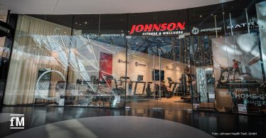 Frontale Ansicht einer Glasfront im MyZeil mit rotem Johnson Fitness & Wellness Logo oben mittig, BowFlex, Vision Fitness, Schwinn, Horizon Fitness und Matrix Fitness Logos auf der Scheibe, dahinter Laufbänder und weitere Fitnessgeräte im Innenraum
