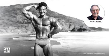 Schwarz-Weiß-Foto eines muskulösen Mannes in Badehose am Strand, der lächelnd den linken Arm anspannt, Meer und Felsen im Hintergrund.