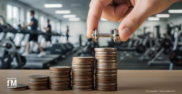 Die Hand hält eine kleine Hantel über gestapelte Münzen auf einem Tisch. Im Hintergrund sind unscharf Fitnessgeräte und trainierende Personen zu erkennen.