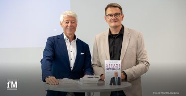 Zwei Männer stehen nebeneinander an einem Stehtisch. Links Prof. Dr. Thomas Wessinghage im blauen Anzug, rechts ein Moderator im beigen Sakko. Beide halten das Buch 'Lebenselixier Bewegung' in der Hand.