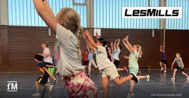 Kinder in einer Turnhalle beim Born-to-Move-Programm von Les Mills, im Vordergrund ein Mädchen mit Zopf und bunter Hose, das springt; im Hintergrund weitere Kinder in Bewegung. Recht oben ist das Les Mills Logo zu sehen.