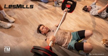 Ein Mann liegt auf einem hellen Parkettboden und drückt eine Langhantel nach oben, weitere Personen sind neben ihm zu erahnen, links oben das LES MILLS Logo in schwarz