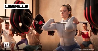 Person steht im Fitnesskurs und hält eine Langhantel in Frontposition, weitere Teilnehmende im Hintergrund.