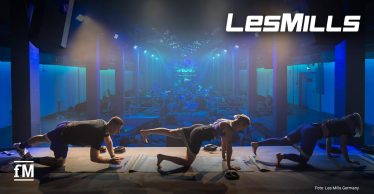 Teilnehmende trainieren auf Matten in einer Halle mit blauer Beleuchtung, im Vordergrund vier Personen in Bodenübungen, im Hintergrund weitere Trainierende. Oben rechts das Les Mills Logo.