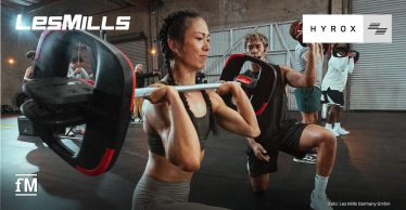 Frau im Vordergrund führt einen Ausfallschritt mit Langhantel aus, im Hintergrund mehrere trainierende Personen in einer Industriehalle.