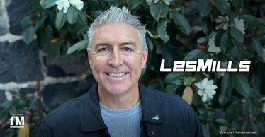 Jason Paris vor grüner Bepflanzung, daneben das weiße Logo von Les Mills.