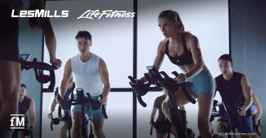 Vordergrund rechts eine Frau auf einem stationären Indoor-Cycling-Bike mit geneigtem Oberkörper und Händen am Lenker, links daneben ein Mann auf einem weiteren Bike; im Hintergrund mehrere Personen auf Fahrrädern in einem abgedunkelten Raum mit großen, hellen Wandflächen