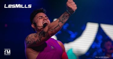 Les Mills Instruktor Sebastian Jaramillo mit auffälligen Tattoos, lila Muskelshirt und Headset ruft laut in Richtung Publikum, das nicht zu sehen ist, und reckt die Faust nach vorne; bunte Bühnenlichter im Hintergrund erzeugen eine kraftvolle Szenerie, während sein Gesichtsausdruck Leidenschaft und Motivation vermittelt.