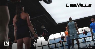 Mehrere Personen trainieren in einem Fitnessstudio mit Langhanteln. Links steht eine Person im Vordergrund mit dem Rücken zur Kamera, rechts trainieren drei Personen vor großen Fenstern. Oben rechts ist das Les Mills Logo zu sehen.