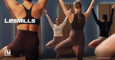 Mehrere Personen stehen barfuß auf Yogamatten in einem Kursraum und balancieren auf einem Bein mit erhobenen Armen. Links etwas oberhalb der Mitte ein weißes Les Mills-Logo. Links unten in der Ecke, das fM Icon, ebenfalls in Weiß.