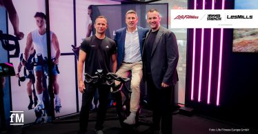 Im Vordergrund stehen zwei Männer neben einem Indoor Bike; ein dritter Mann sitzt auf dem Sattel. Der linke Mann trägt ein schwarzes Shirt mit den Schriftzügen „LES MILLS“ und „adidas“. Links im Hintergrund zeigt eine große Wandgrafik eine Person auf einem Bike, rechts stehen vier vertikale pinke Lichtleisten vor einer dunklen Wand.
