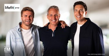 Drei Männer stehen nebeneinander vor hellem Hintergrund, links das Logo der „Lifefit Group“, unten links das fM-Logo, unten rechts eine Bildquelle „Foto: LifeFit Group MidCo GmbH“.