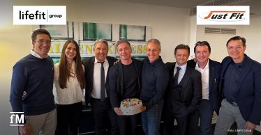 Gruppenaufnahme in einem Innenraum in Köln mit mehreren Personen aus dem Management von Just Fit und der LifeFit Group, Logos beider Unternehmen im oberen Bildbereich.